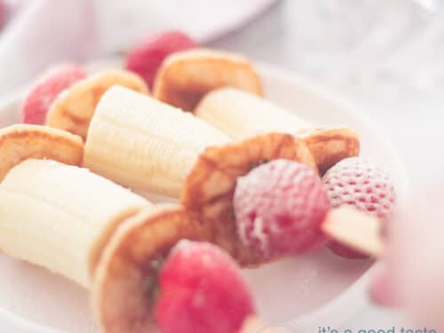 A square photo with three sticks filled with banana, pancakes and strawberries. A hand takes one off the board.