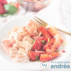 A square photo with a white plate filled with balsamic strawberries, ham, a golden fork and the ingredients in the background