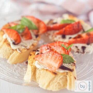 A square picture of a glass plate with three bruschetta with mascarpone, balsamic, strawberries and basil.