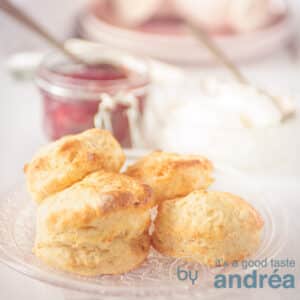 A square photo with a glass plate of scones and a jar of strawberry jam in the background.