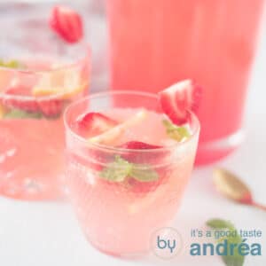 A square photo with two glasses filled with strawberry lemonade with fresh fruit and a jug in the background