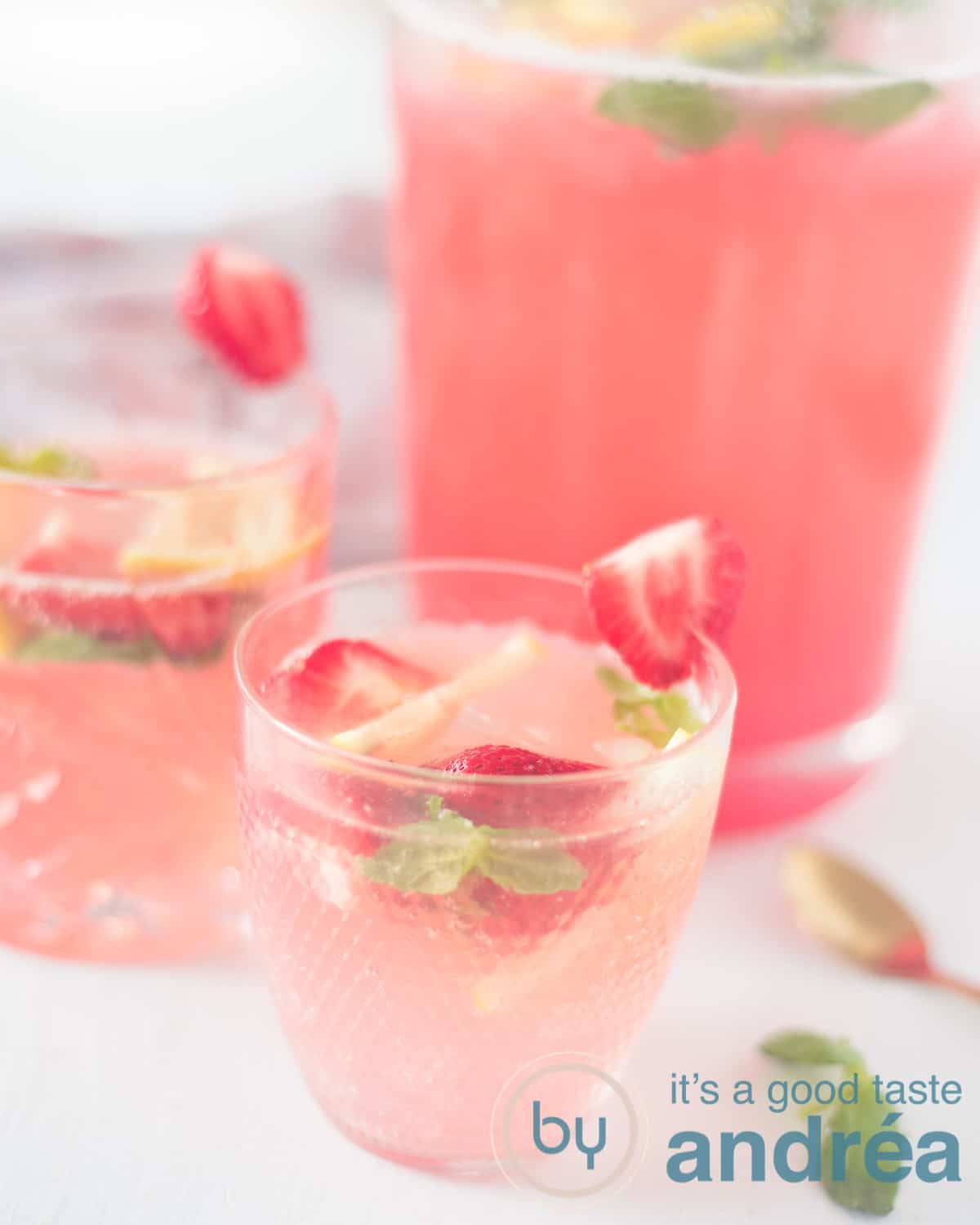 An elevation shot with two glasses of strawberry and basil lemonade and a pitcher with the juice in the background.