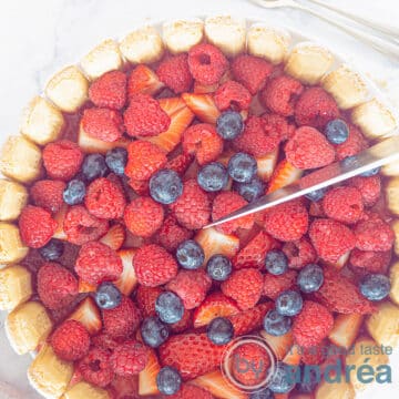 A square photo of a charlotte russe from above. A topping of raspberries, blueberries and strawberries. A knife cuts off a wedge.