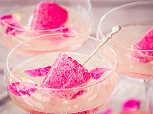 A square photo with three champagne glasses with bubbles, raspberry ice cream collies, and rose petals