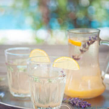 two glasses with lavender lemonade, garnished with lemon. A jar of lemonade and a summer background