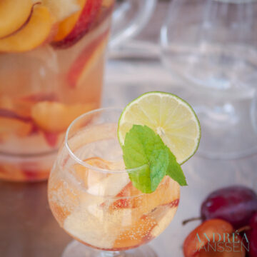 A glass of sangria Bianca with fresh fruit and a mint and lemon garnish. In the back a jar and some glasses