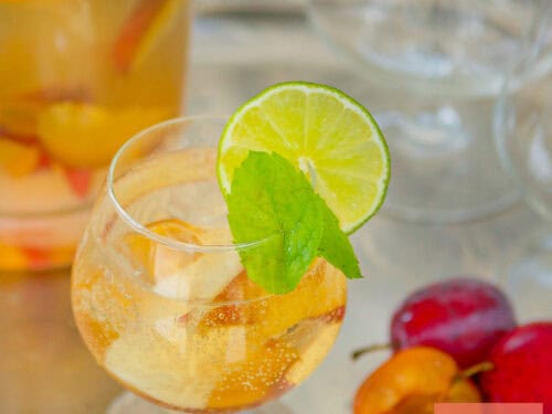 A square photo with a glass of sangria with cava and summer fruit. A pitcher behind it.