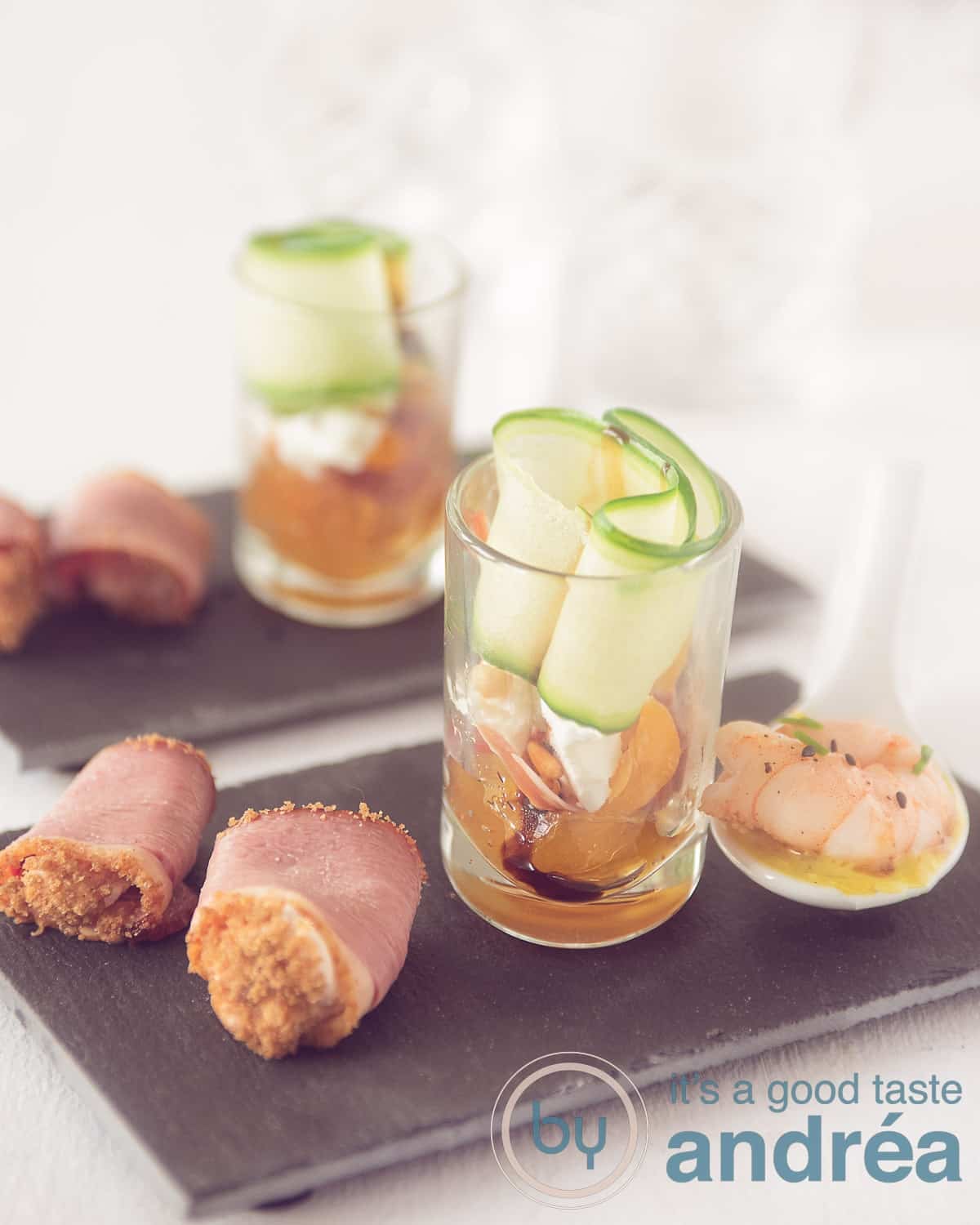 An elevation photo with two times three amuses on a black chalkboard. A spoonful with shrimp, a glass with cucumber and a roll of duck breast.