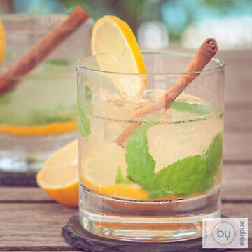 A square picture with two glasses filled with ginger caipirinha and some mint leaves, lemon and cinnamon