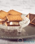 Three s'mores with chocolate biscuits on a glass plate. Some chocolate cookies in the back