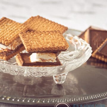 Three s'mores with chocolate biscuits on a glass plate. Some chocolate cookies in the back