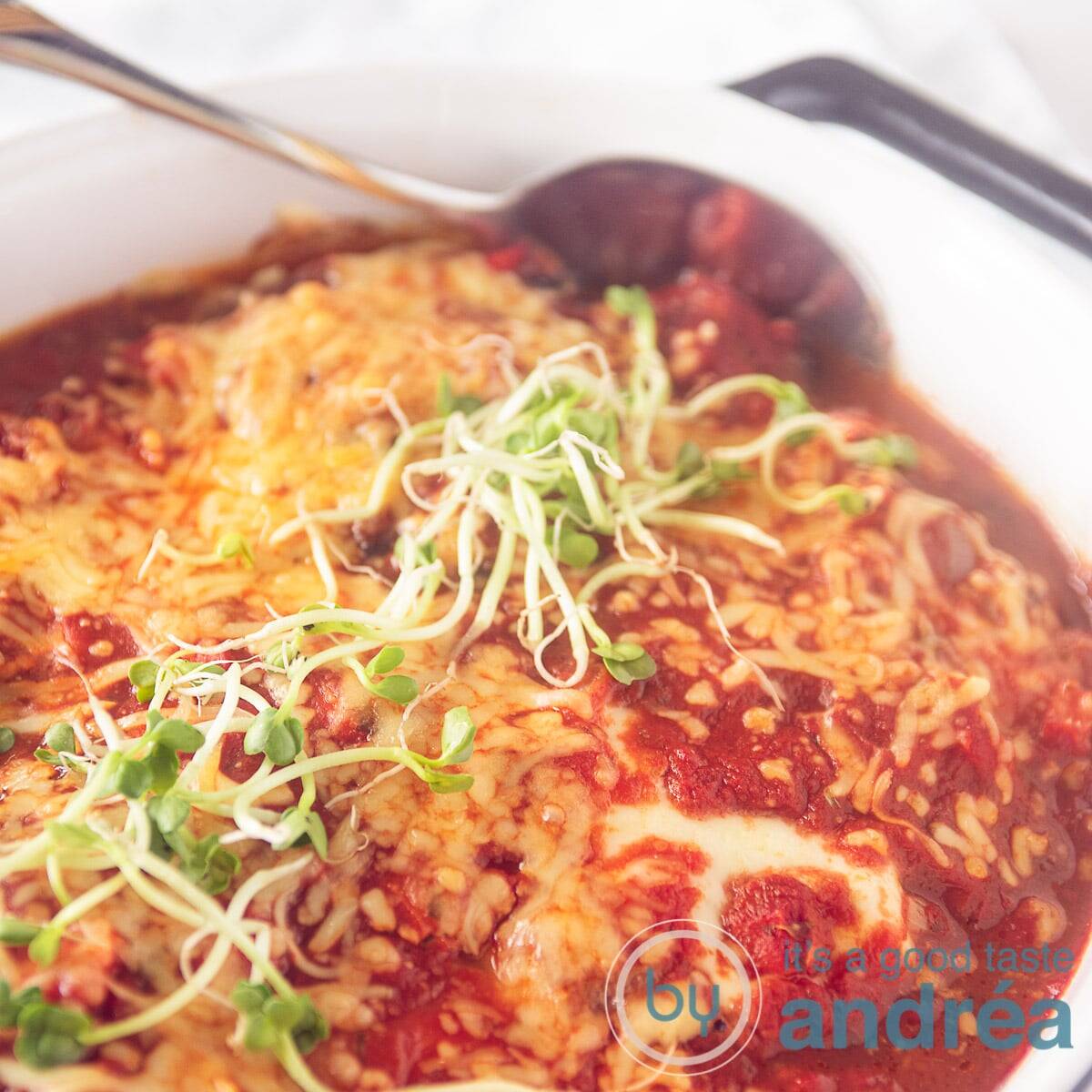 A square photo with half a baking dish filled with tomato sauce, schnitzel and cheese. A spoon sticks in the oven dish