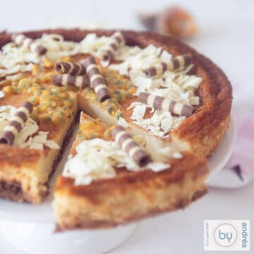 Een vierkante foto van een cheesecake met witte chocola en passievruchten. Een topping van vlokken en slagroom.