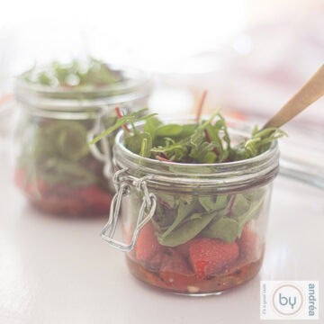 A square picture with two jars filled with fresh salad with lettuce, strawberries and honey mustard dressing