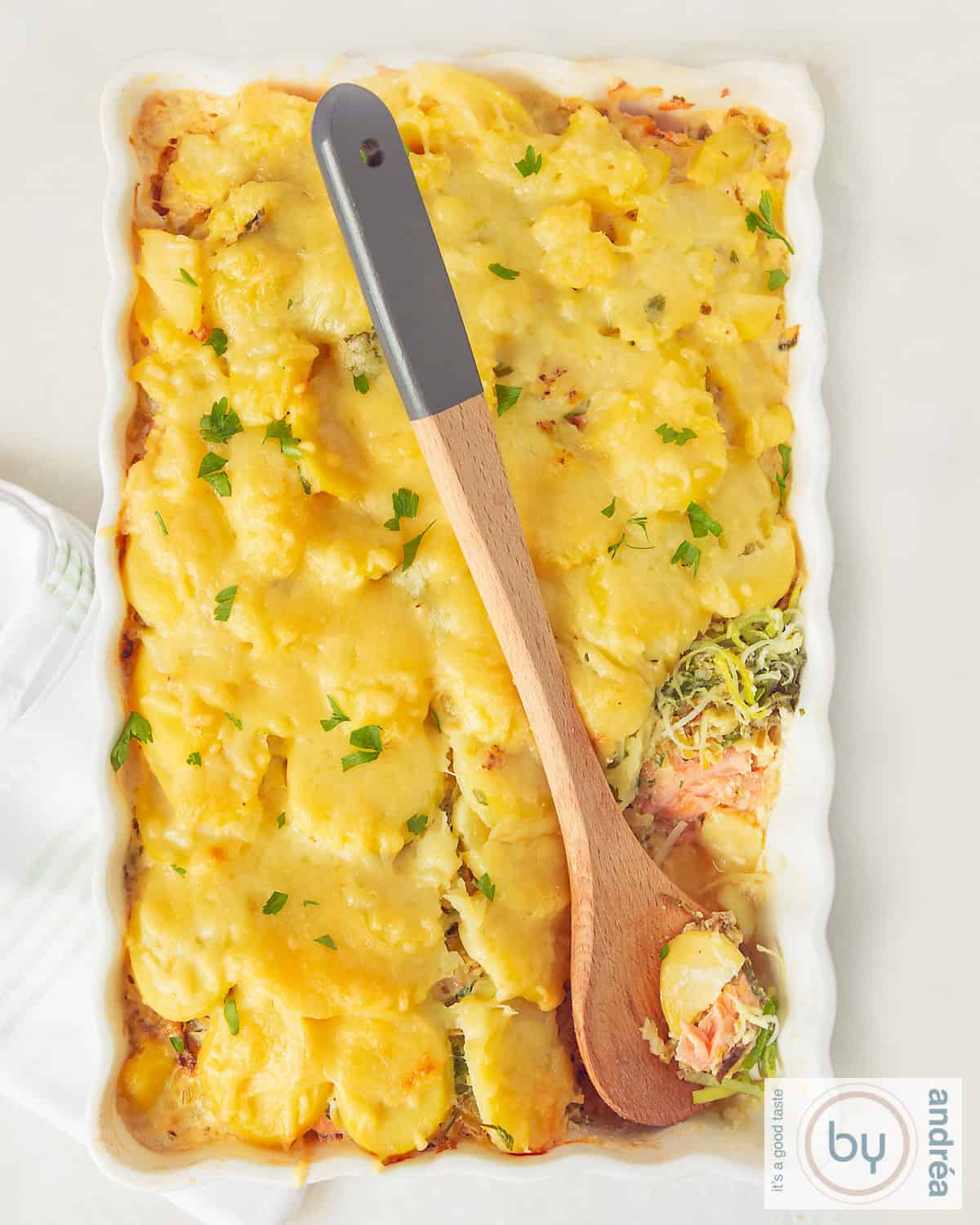A photo of a white casserole dish with salmon, leek, and potato casserole with cheese. A wooden spoon is placed in the casserole dish.