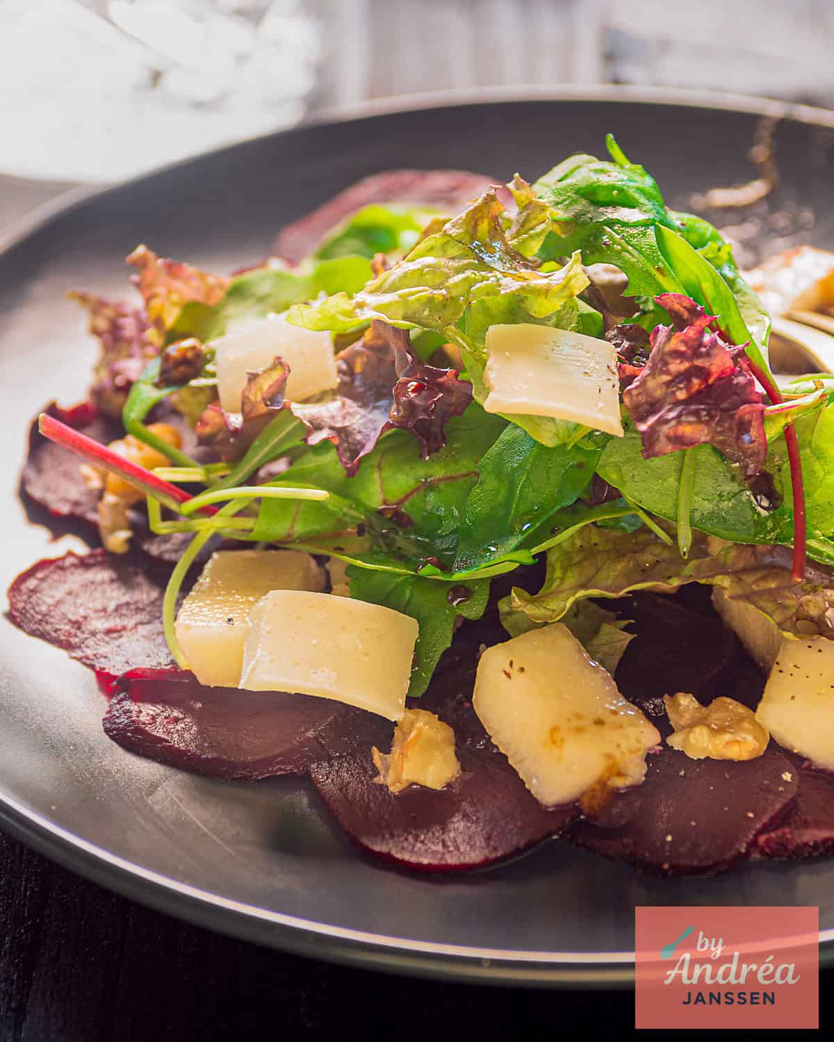 Een foto van een deel van een grijs bord met bietencarpaccio met kaas, walnoten, sla en peer. 