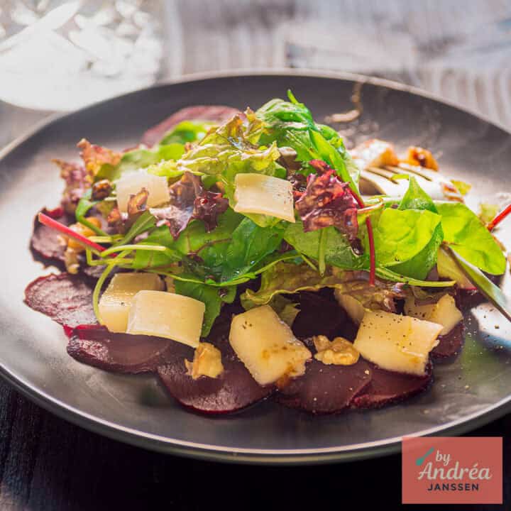 A square photo with a plate of beetroot, pear, cheese, lettuce, and walnut carpaccio.