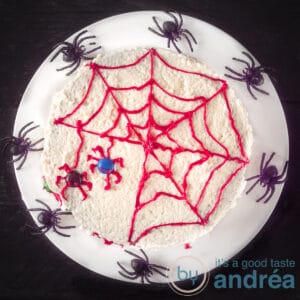 A square photo with a white plate with a red spider web cake and two spiders. Spin around it.