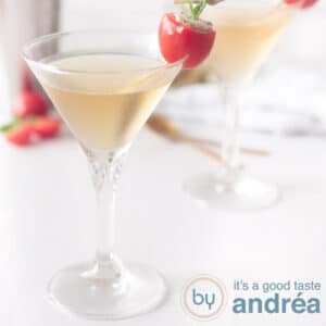 A square photo with two cocktail glasses filled with a basil martini cocktail. On a white background a cherry tomato as garnish
