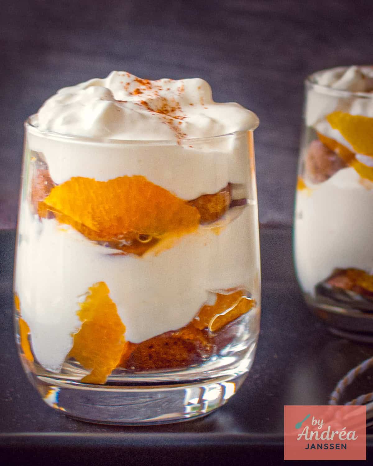 Twee glazen gevuld met tiramisu met pepernoten en mandarijn op een zwarte ondergrond.