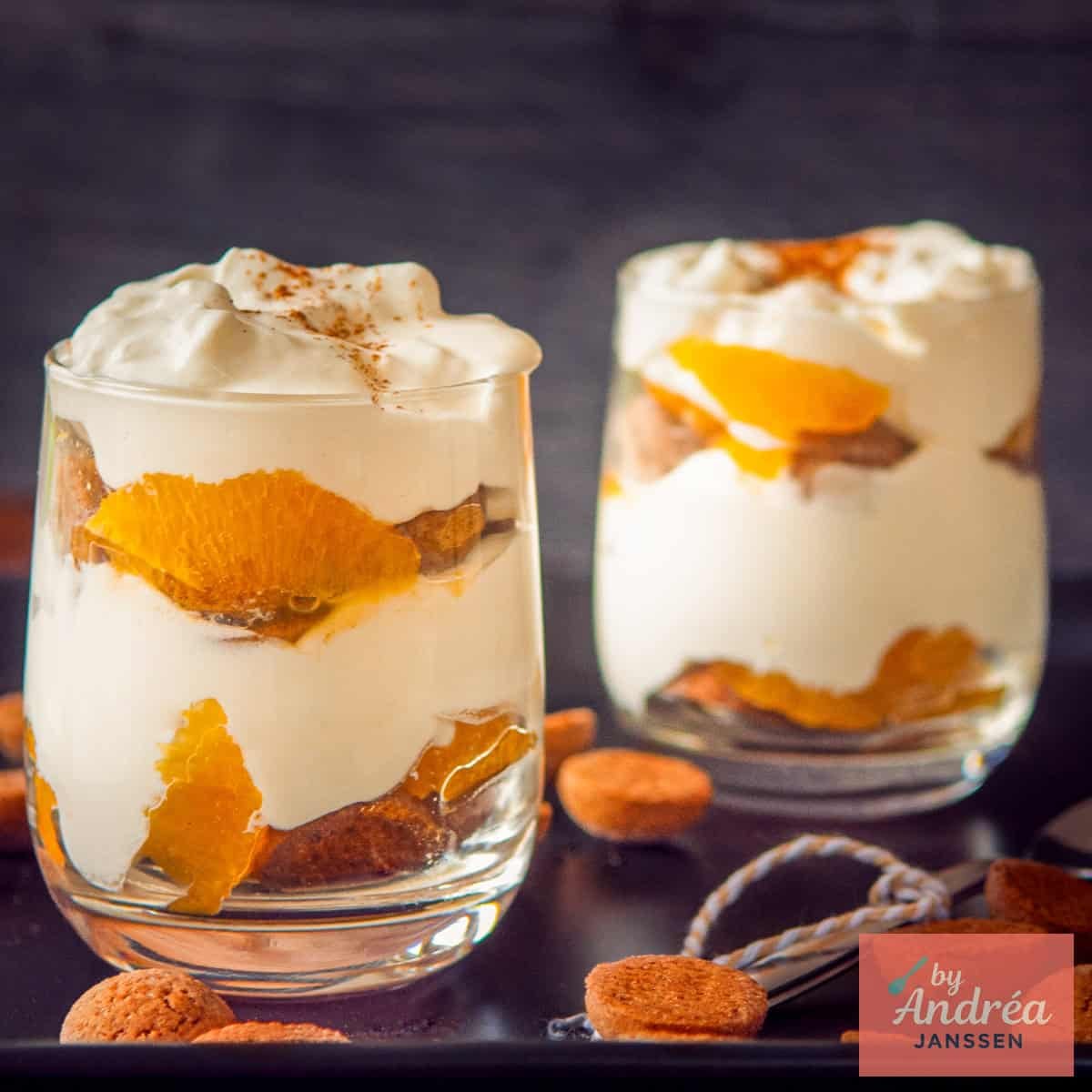 A square photo with two glasses of pepernoten mandarin tiramisu diagonally behind each other on a black background.