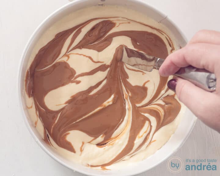 A springform pan with cheesecake filling into which speculoos has been scooped. The back of a knife is swirled through the batter.