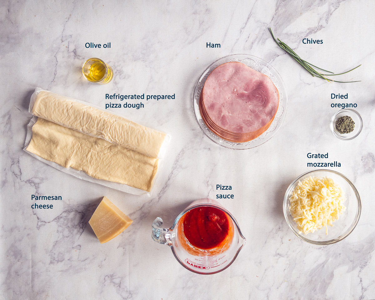 Labeled ingredients for mini Christmas pizzas: pizza dough, pizza sauce, ham, grated mozzarella, chives, Parmesan cheese and olive oil on a marble background