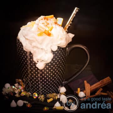 A square photo of a black mug with whipped cream, golden chocolate curls and cinnamon marshmallows on the black background.