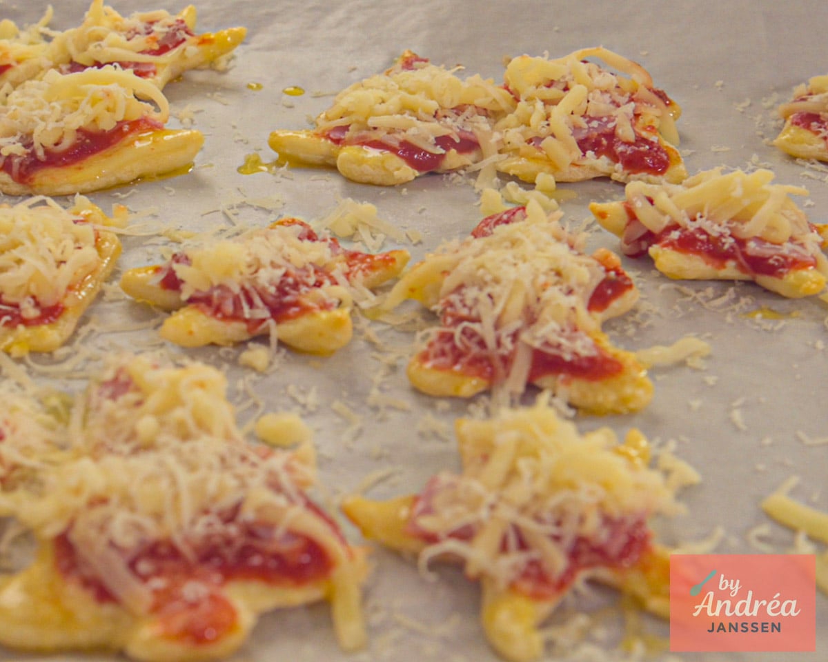 Ongebakken mini pizzabodems in kerstvormen belegd met pizzasaus en geraspte kaas op bakpapier