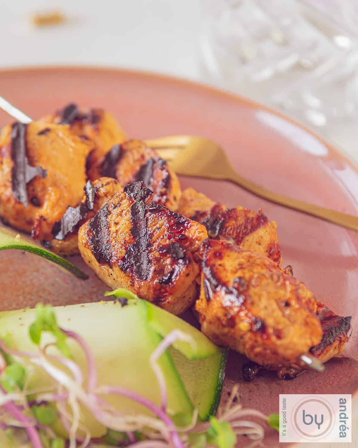 An aerial photo of two chicken skewers marinated in ketjap and mustard on a pink plate. A cucumber salad is served with it.