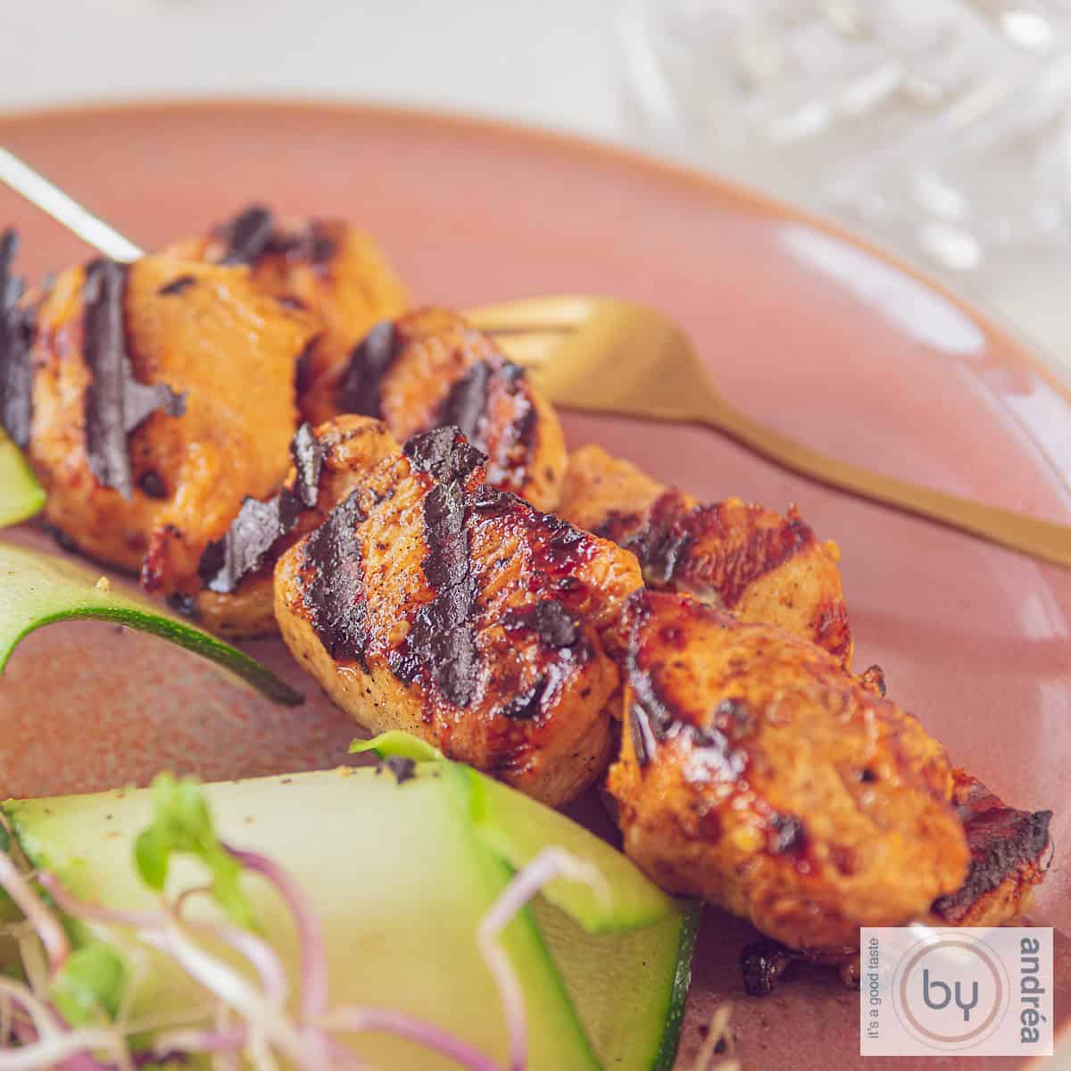 A square photo of two skewers on a red plate with chicken marinated in mustard and ketjap and grilled.