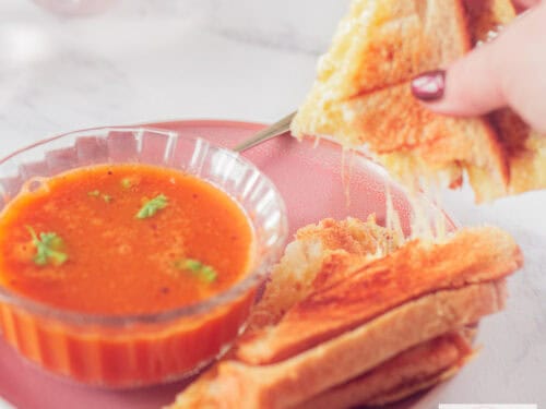 A square photo of a bowl of tomato soup with parsley and a grilled cheese sandwich with a piece broken off, revealing streaks of melted cheese