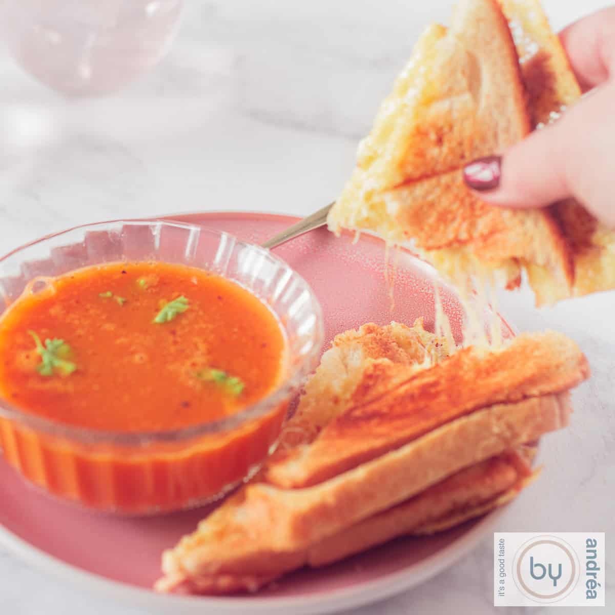A square photo of a bowl of tomato soup with parsley and a grilled cheese sandwich with a piece broken off, revealing streaks of melted cheese
