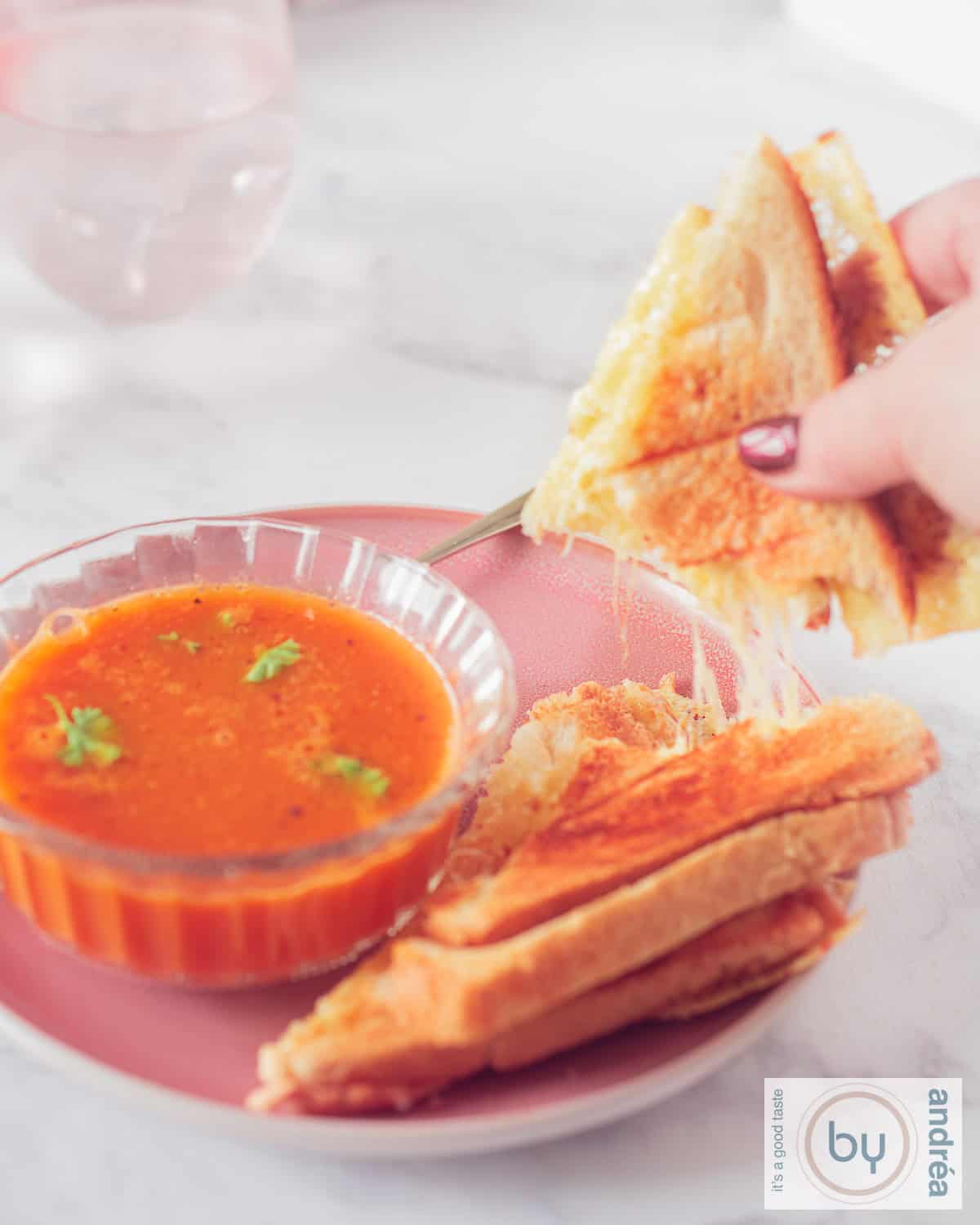 A photo of a pink plate with a glass bowl of tomato soup and a grilled cheese sandwich with a piece torn off, revealing strings of cheese.