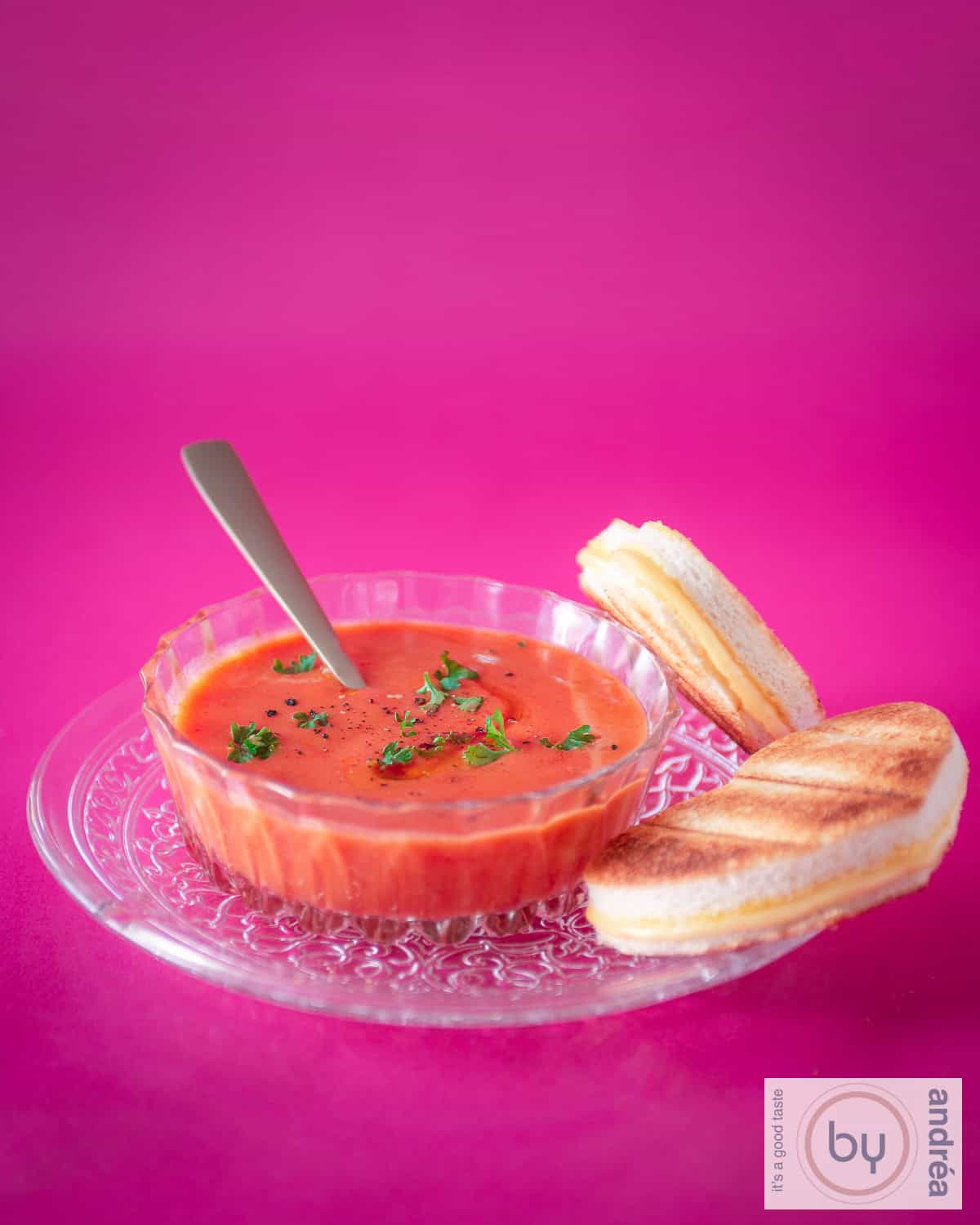 A height photo with a pink background, a glass plate with tomato soup in a glass bowl and heart-shaped toasted sandwiches