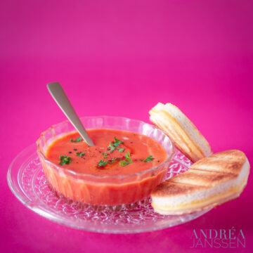 a glass bowl with tomato soup and grilled cheese sandwiches on a pink background