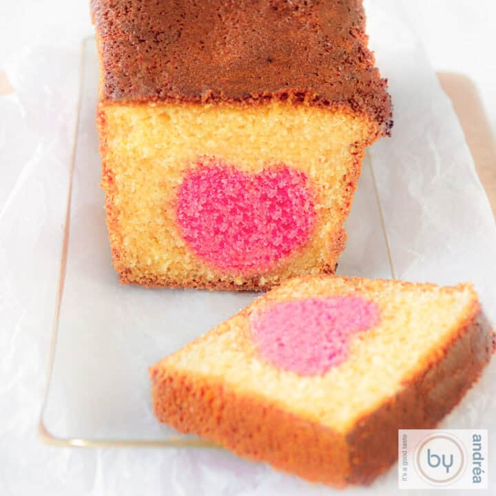 Een vierkante foto met een glazen schaal met daarop een cake waar een plakje is afgesneden. Een roze hart kun je zien