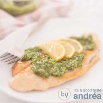 A square photograph showing part of a white plate with arugula pesto topped pangasius fillet with lemon slices.