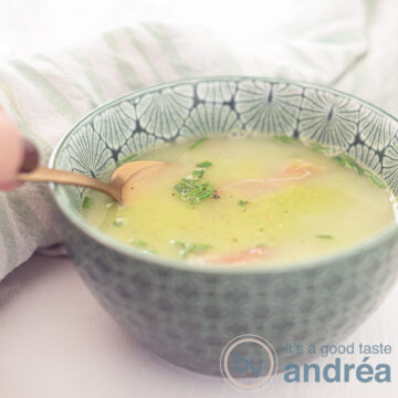 A square photo with a green bowl filled with green pea soup, Serrano ham, cream and chives. A hand takes a bite. A white background.