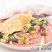 A square photo with a pink plate in the foreground, with a filled omelette with arugula, spring onions, shrimps and ham. In the background a pink, white tea towel.