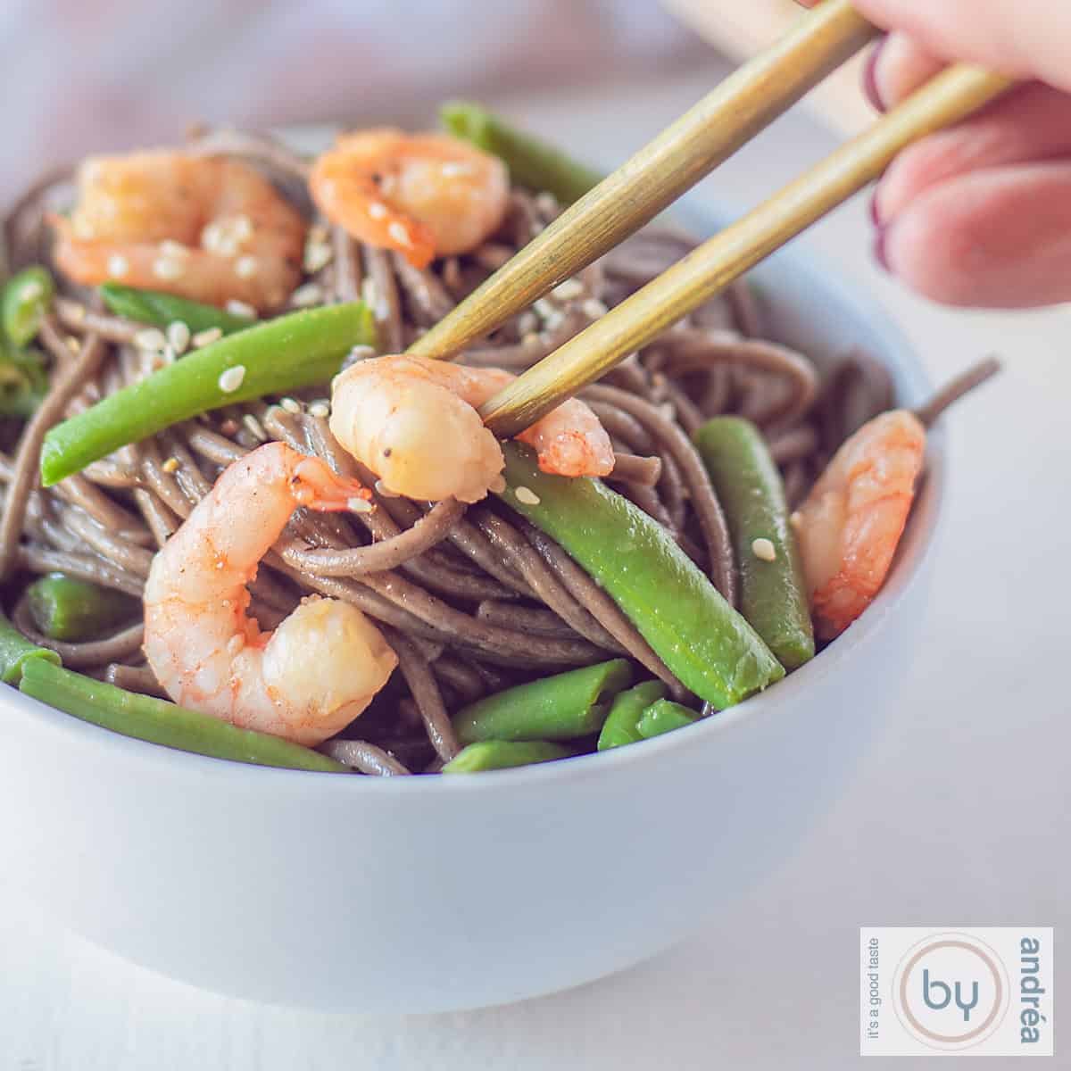 A square photo of a white bowl filled with soba noodles with shrimp, green beans, and sesame seeds. Two chopsticks pick up a shrimp from the bowl.