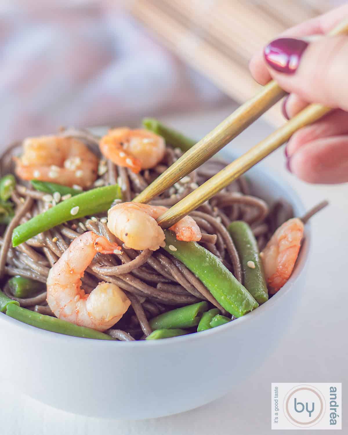 Een deel van een wit schaaltje waar twee chopsticks een garnaal pakken uit een bakje gevuld met garnalen, sperziebonen, noedels en sesam
