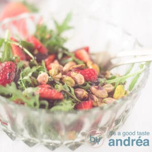 A square photo with part of a glass bowl filled with arugula, strawberries and pistachio nuts