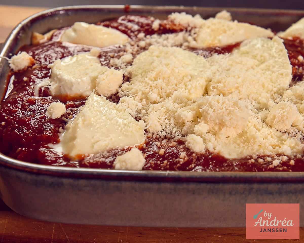 Baking dish with chicken and tomato sauce, topped with sliced mozzarella and grated Parmesan cheese.