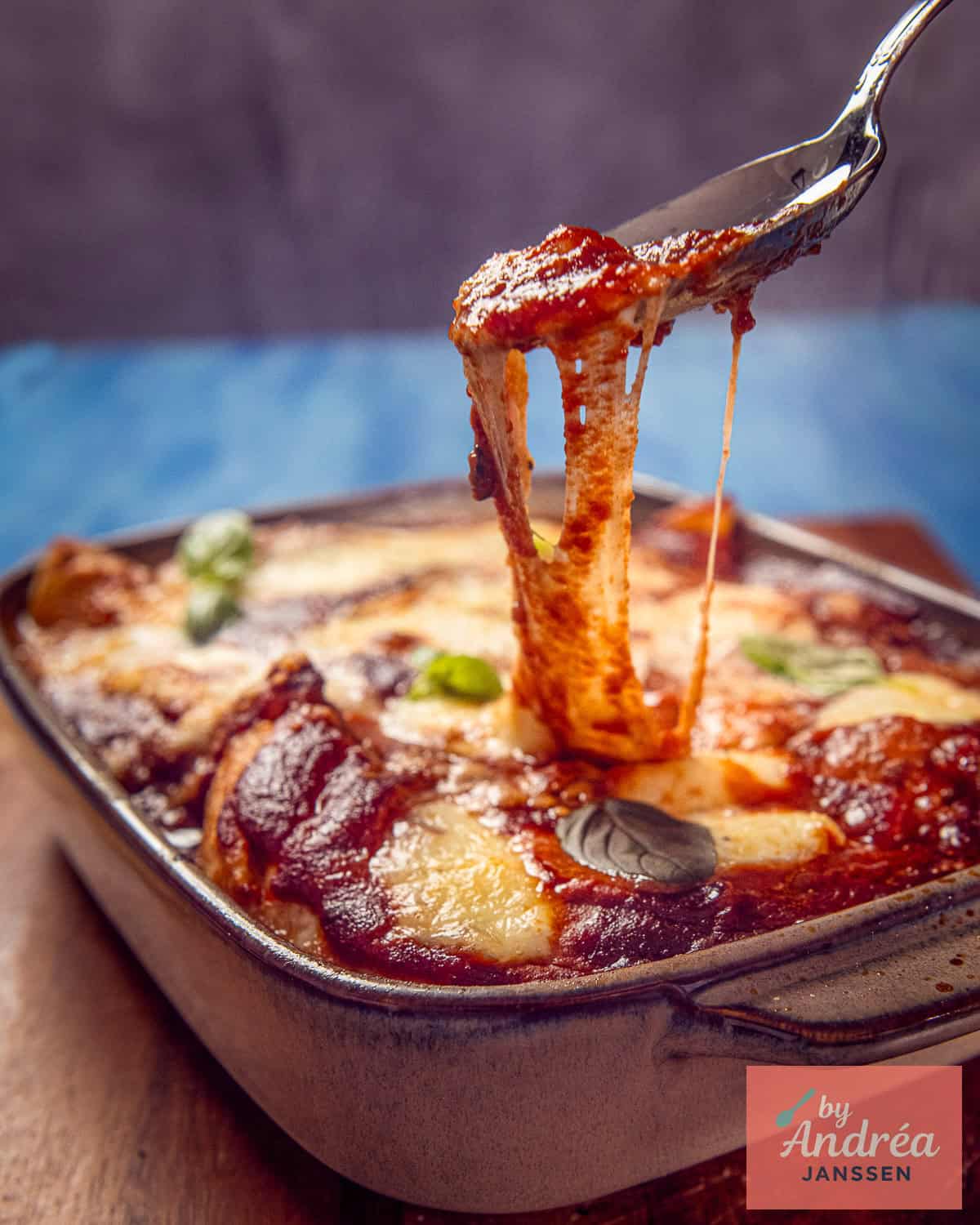 A piece of chicken pizzaiola is lifted from the baking dish, with long strands of melted mozzarella.
