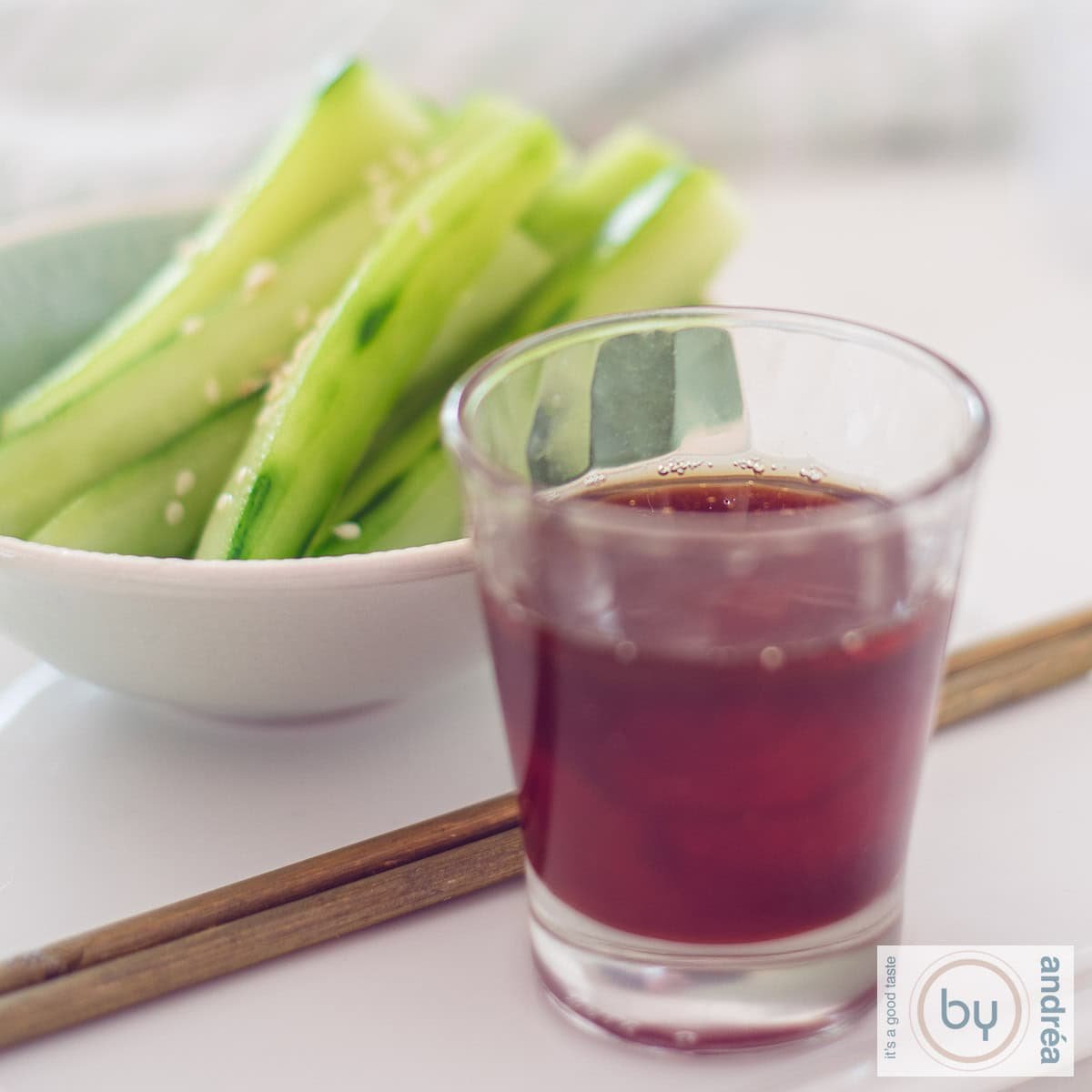 A square photo with a bowl of cucumber and sesame strips in the background, two chopsticks and a glass with a spicy dipping sauce in the front.