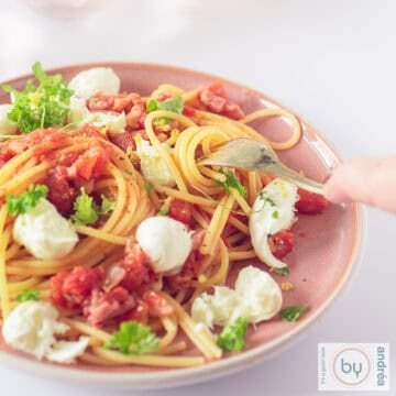 A square photo of a pink plate filled with spaghetti, bacon, tomatoes, mozzarella and parsley