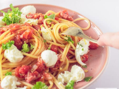 A square photo of a pink plate filled with spaghetti, bacon, tomatoes, mozzarella and parsley