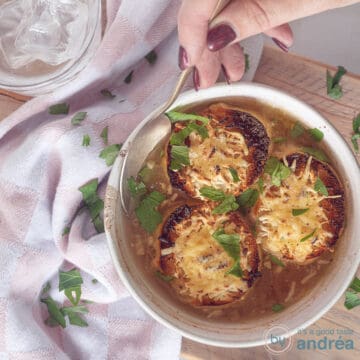 A photo from above of a white bowl of onion soup with three pieces of gratinated baguette. A spoon takes a bite. A pink and white tea towel is next to it on the left.
