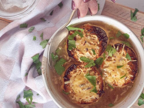A photo from above of a white bowl of onion soup with three pieces of gratinated baguette. A spoon takes a bite. A pink and white tea towel is next to it on the left.